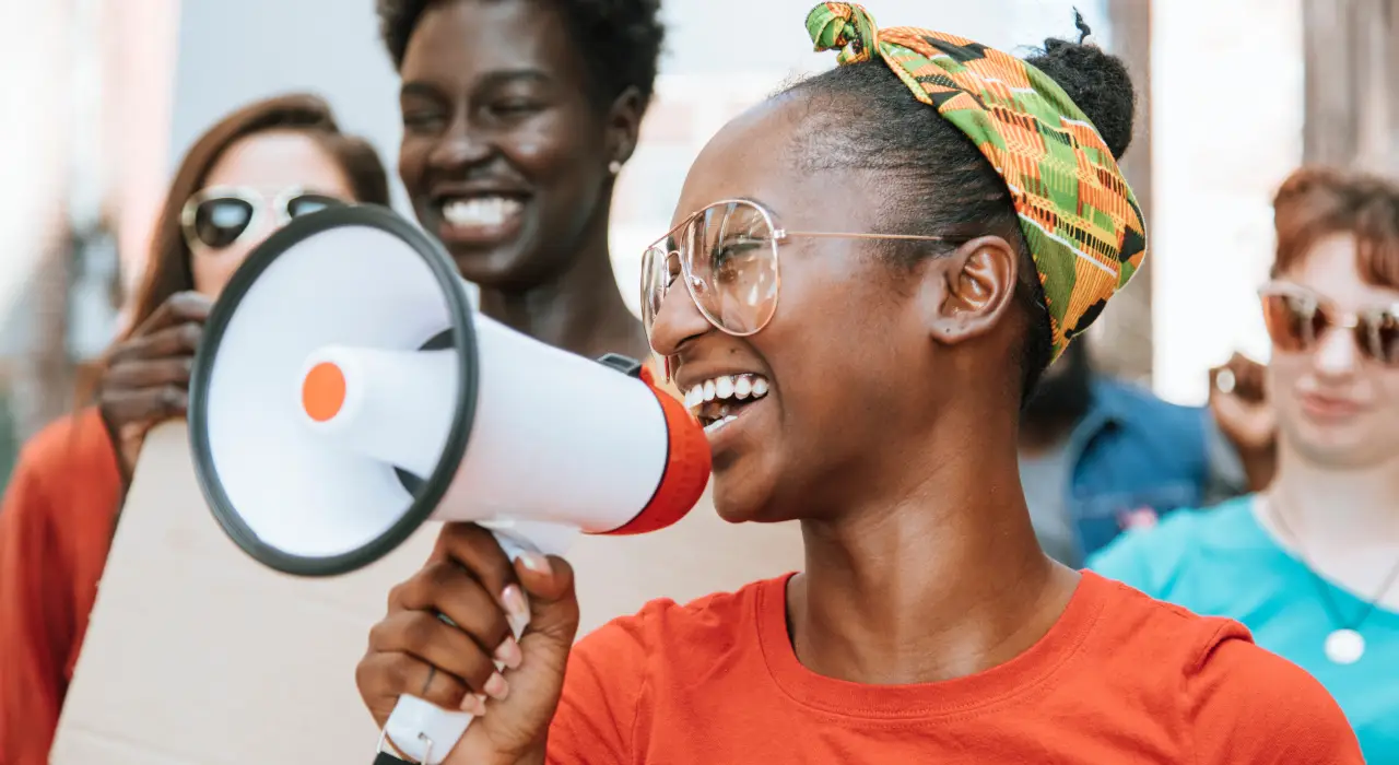 ਇੱਕ ਔਰਤ ਬਾਹਰਲੀ ਰੈਲੀ ਵਿੱਚ ਮਾਈਕ ਰਾਹੀਂ ਬੋਲ ਰਹੀ ਹੈ, ਉਸ ਦੇ ਪਿੱਛੇ ਹੋਰ ਲੋਕ ਹਨ।