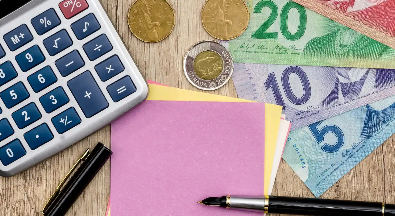 Una calculadora, monedas y billetes canadienses sobre un escritorio junto a un bolígrafo y notas adhesivas.