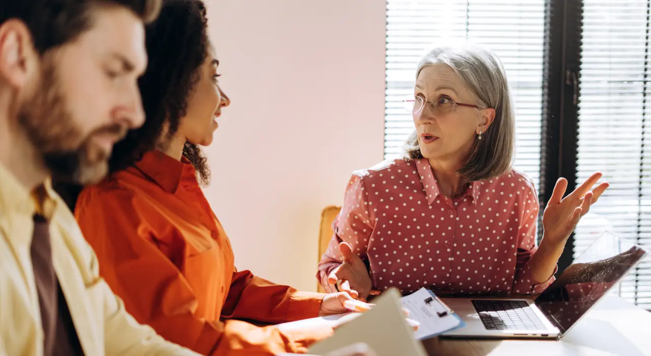 Tres personas sentadas en una mesa con portátiles y documentos, en una discusión enfocada.