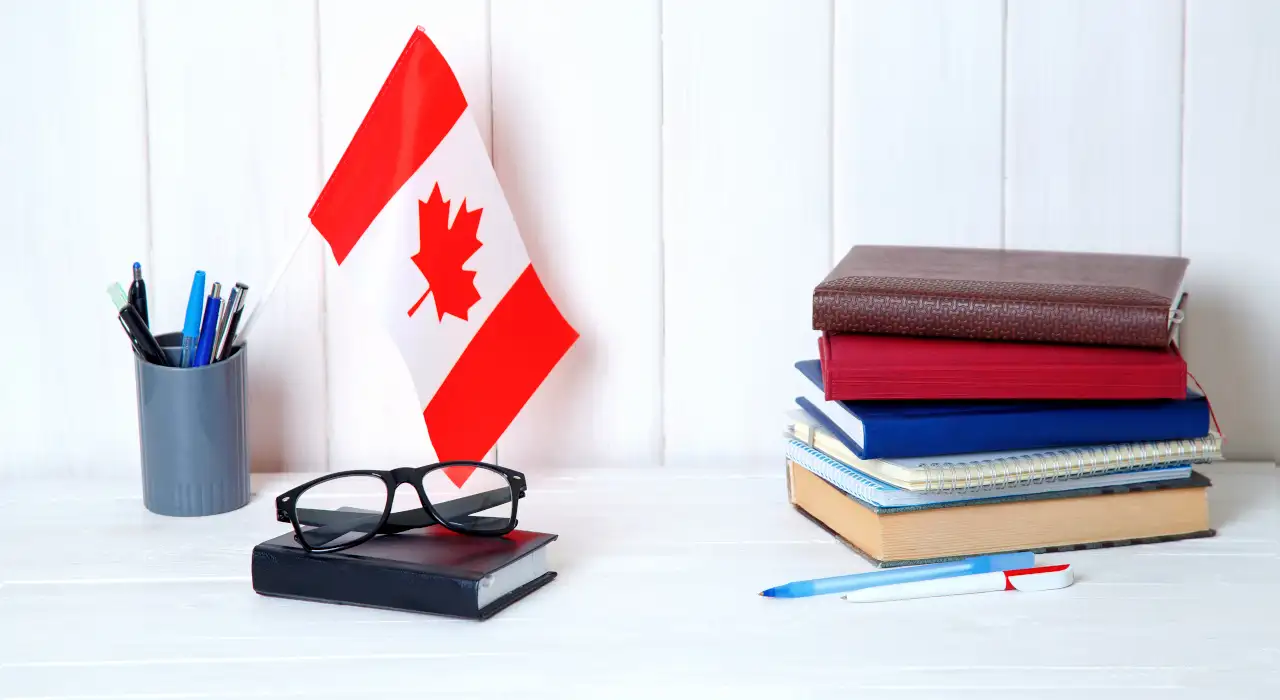 Una pila de libros y cuadernos junto a un portalápices, unas gafas y una pequeña bandera canadiense en un escritorio.