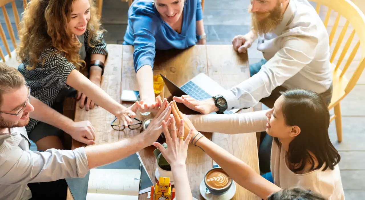 六人圍坐在木桌旁，笑著把手疊在一起作團隊手勢。