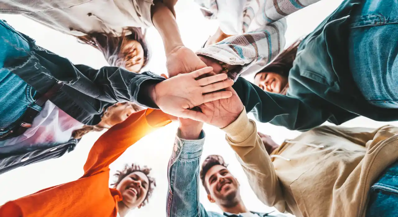 Un grupo diverso de jóvenes sonríe mientras forman un círculo, apilando sus manos juntas en señal de unidad.